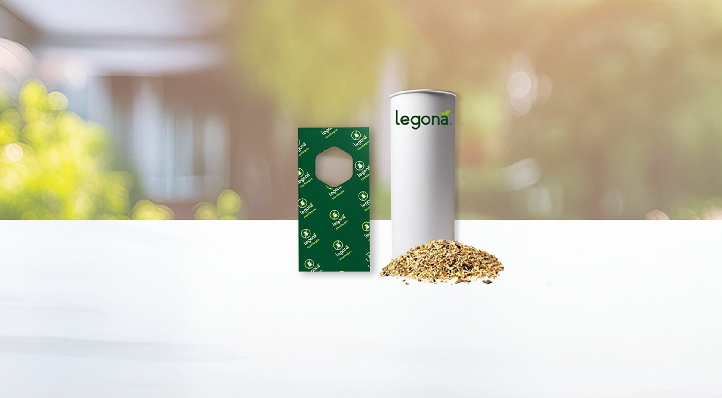 Zylindrische weiße Verpackung mit grünem Schriftzug "legona" neben grünem Trägerstreifen und einem Haufen Granulat auf weißer Oberfläche vor unscharfem Garten-Hintergrund