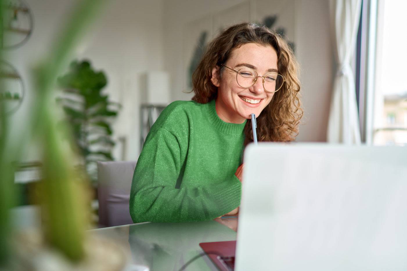 Junge Frau mit Brille und grünem Pullover lächelt vor einem Laptop und hält einen Stift