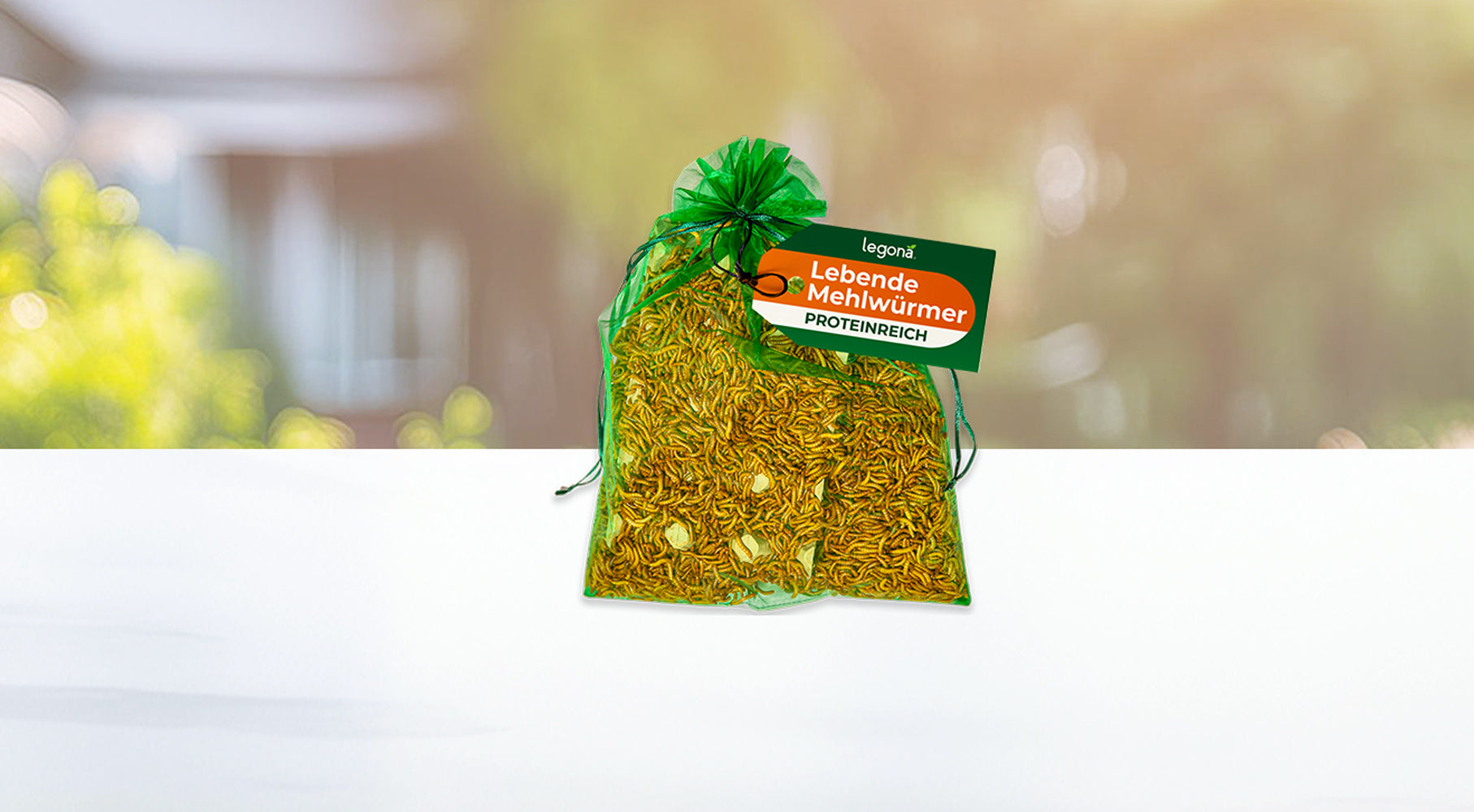 Grüner Netzbeutel mit lebenden Mehlwürmern und grün-orangefarbenem Etikett mit der Aufschrift 'Lebende Mehlwürmer proteinreich' auf einem weißen Tisch, vor unscharfem Gartenhintergrund